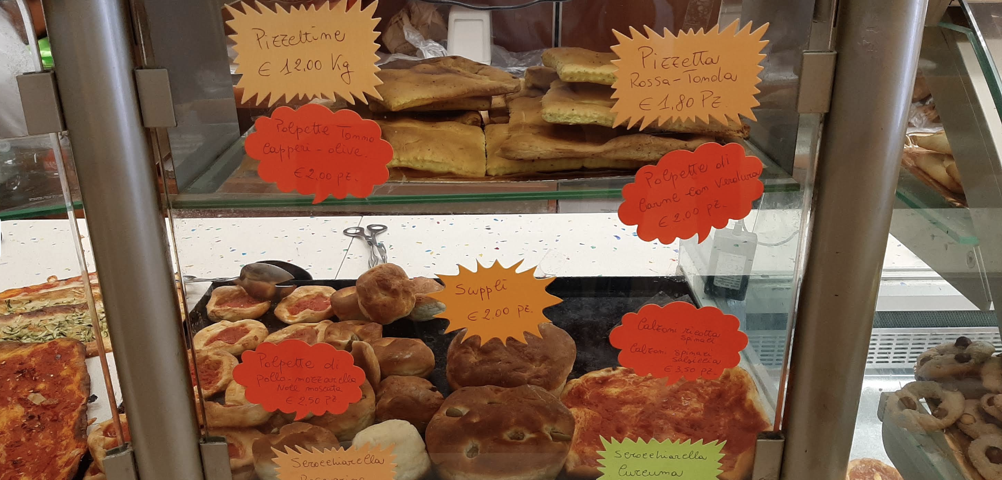 The glass display case at Panificio Monti with focaccia, meatballs, supplì and calzoni, each with a handwritten label