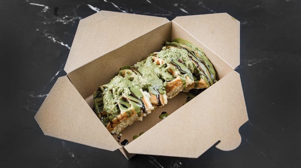 A waffle topped with pistachio cream and chocolate drizzle in a paper box